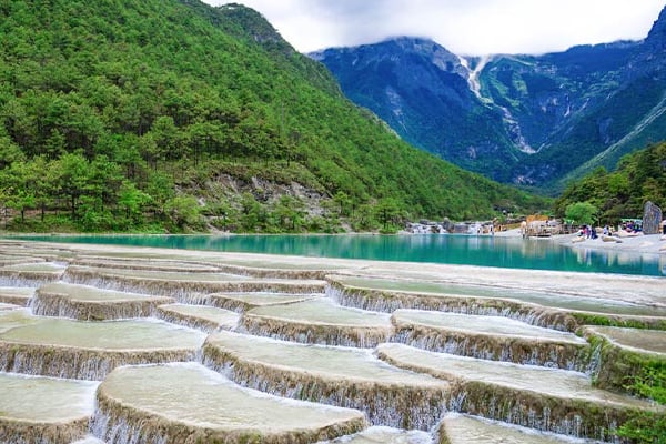 Terraced travertine pools of Blue Moon Valley with turquoise water, forested slopes and snow mountains, Yunnan